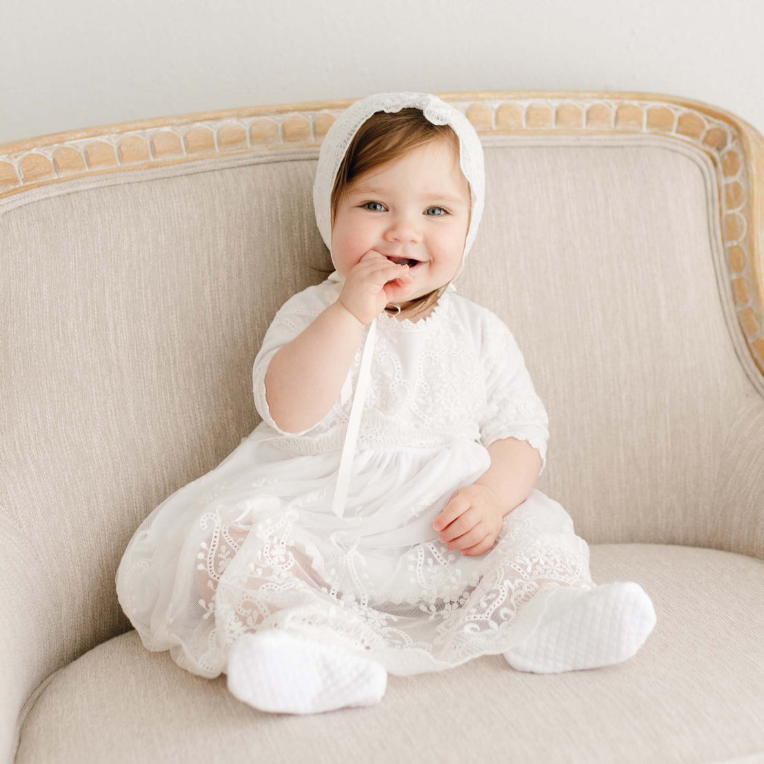 A smiling baby in the Eliza Lace Dress & Headband sits on a beige sofa, looking at the camera.