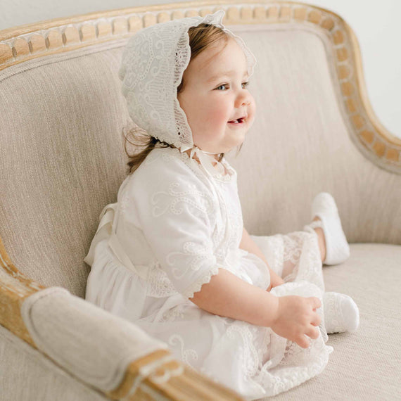 A toddler girl wearing the Eliza Lace Dress & Headband sits on an elegantly upholstered sofa, looking away with a joyful expression. The setting is upscale and serene, with a gentle light illuminating the scene.