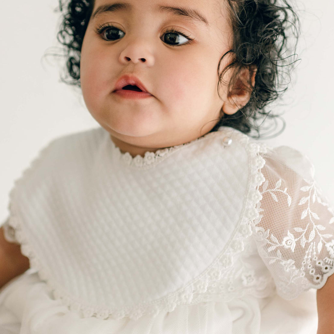 A baby with curly hair and expressive eyes wearing a white outfit with lace detailing and a vintage touch, looking slightly off-camera. The attire includes the Ella Bib made of quilted cotton and short sleeves featuring an intricate floral lace pattern. The background is plain and light.