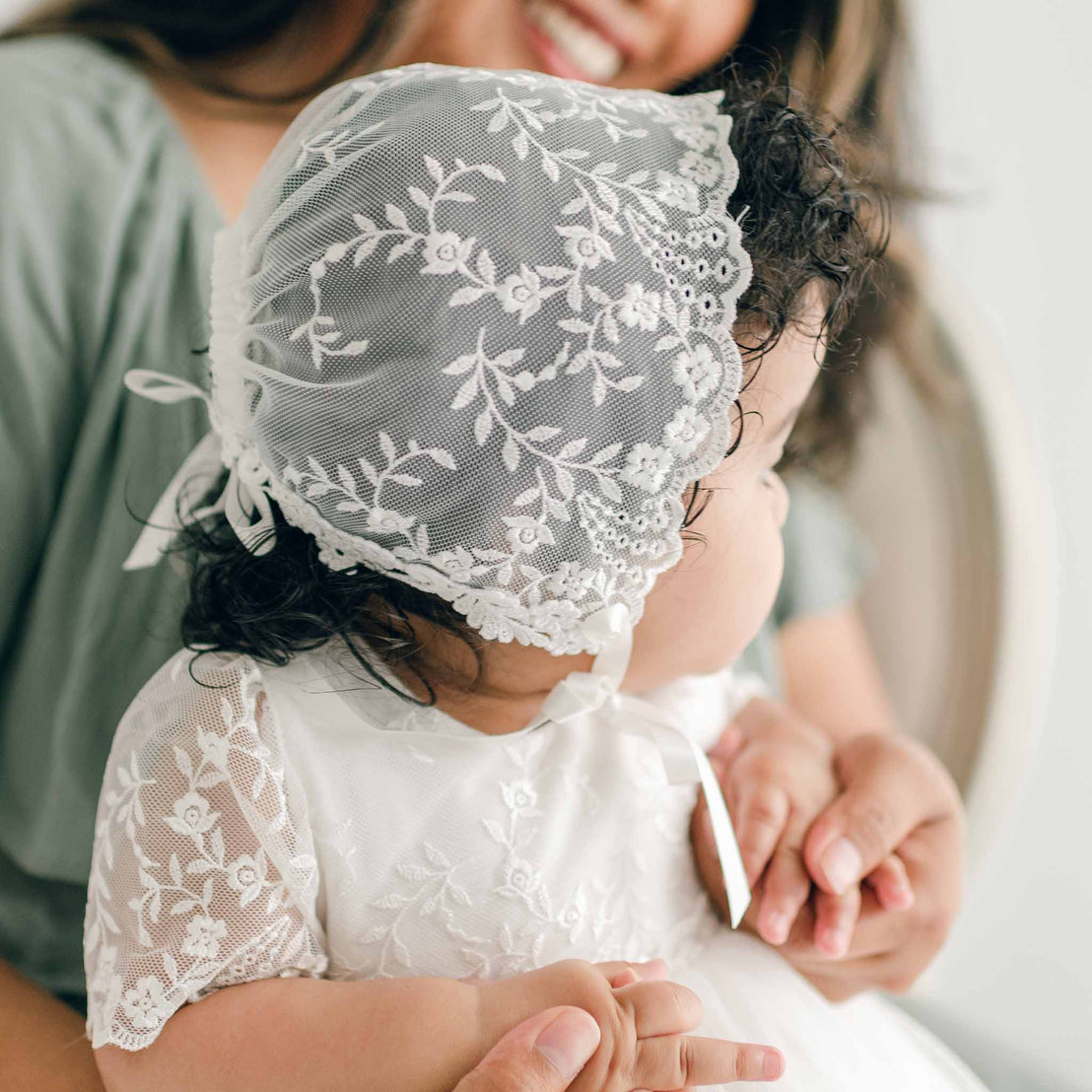 A baby wearing the Ella Lace Bonnet and the Ella Christening Gown sits in a woman's lap. The baby has curly hair and is looking to the side. The woman, wearing a green top, gently holds the baby's arm.