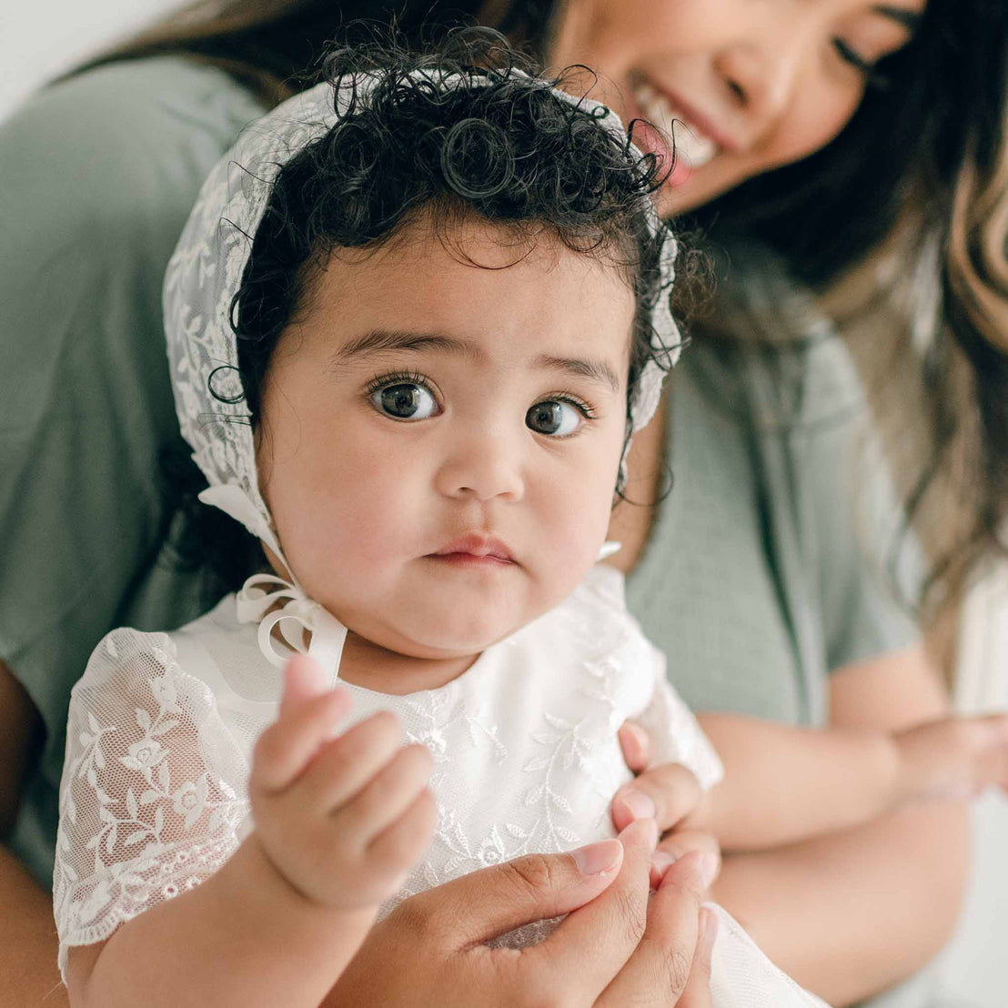 A baby wearing an Ella Lace Bonnet with floral embroidered lace and the Ella Christening Gown gazes into the camera while being held by her smiling mother in the background. The baby has curly hair and large eyes, with a calm expression on their face.