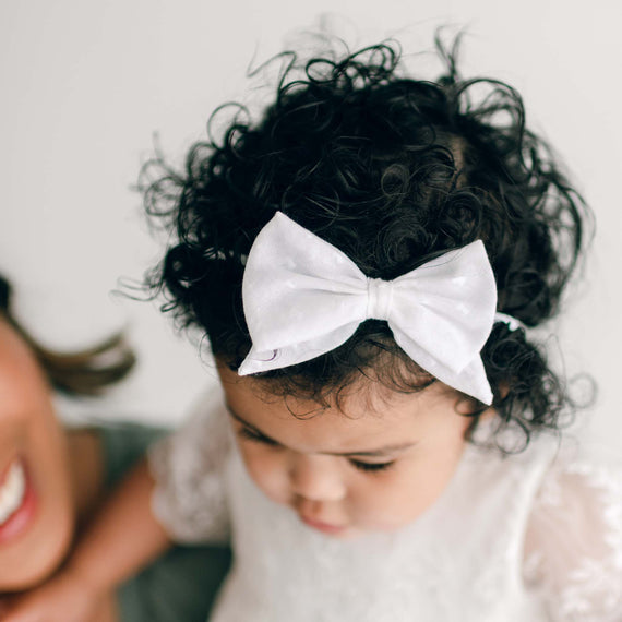 A close-up photo of a toddler with curly hair tied with a white Ella Bow Headband, looking down thoughtfully. The blurred smiling face of an adult is partially visible.