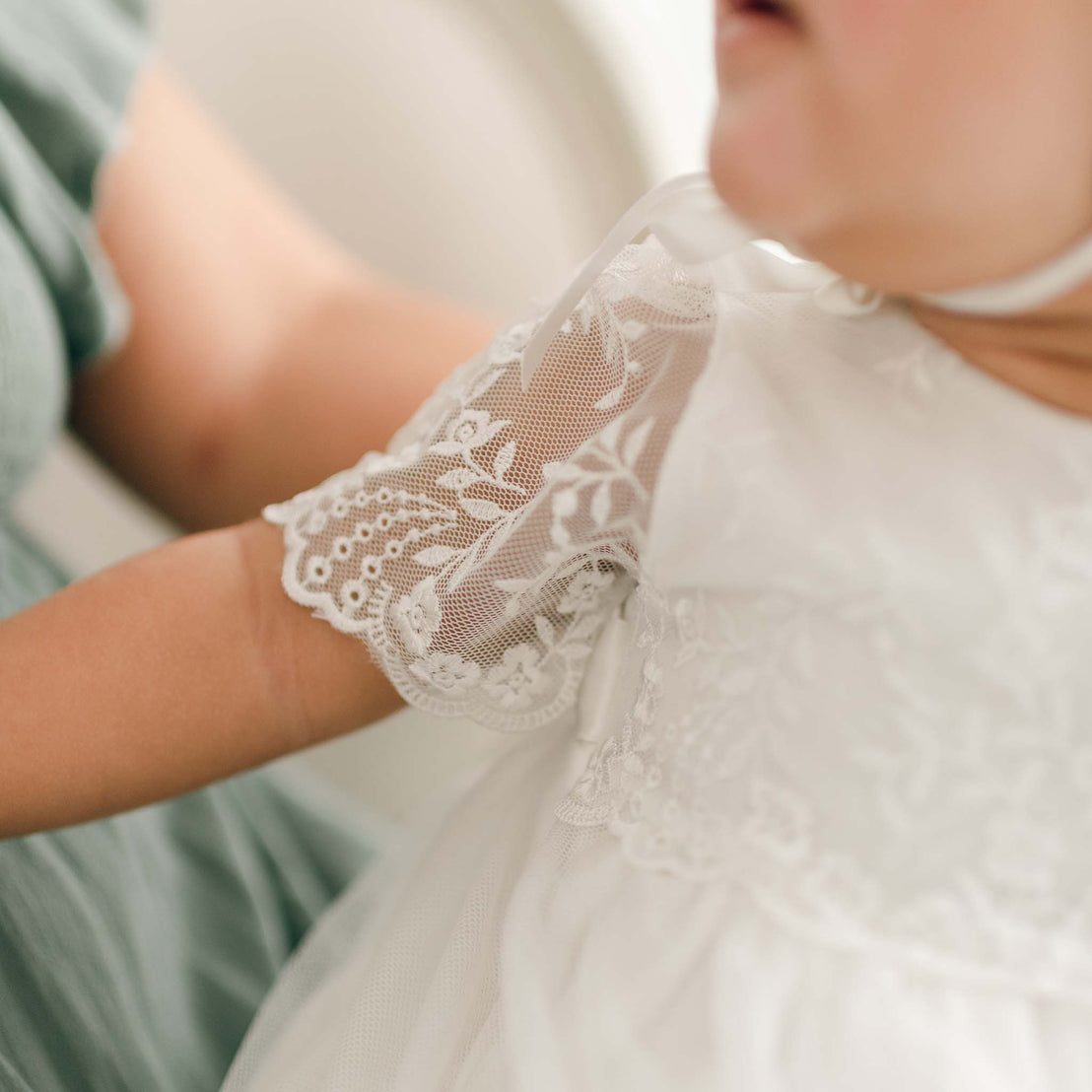 A close-up photo of a baby in an Ella Christening Gown, featuring detailed floral embroidered netting lace sleeves. The intricate lace pattern on the dress sleeve is clearly visible. An adult in a green dress is holding the baby. 