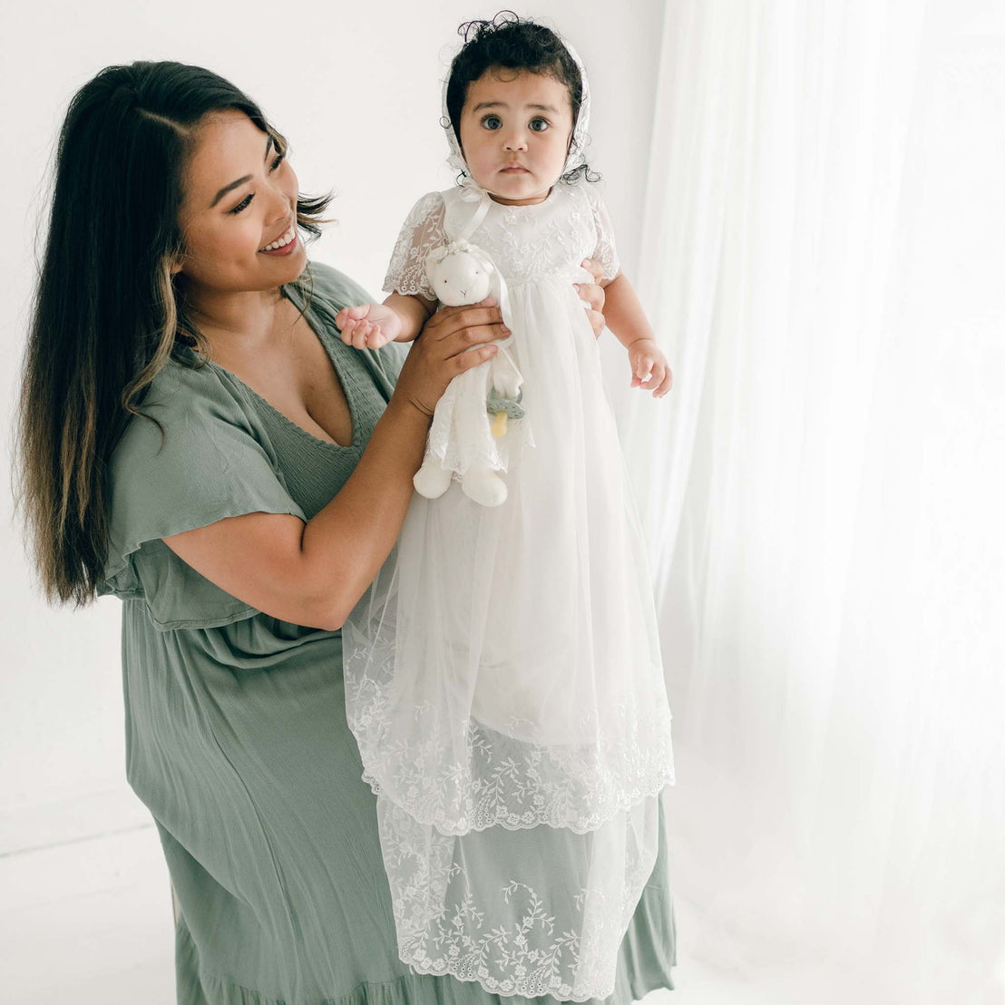 A woman in a green dress smiles while holding her baby girl dressed in the Ella Christening Gown, and a Silly Bunny Buddy. The baby looks at the camera curiously. They are sitting against a soft, white curtain backdrop.