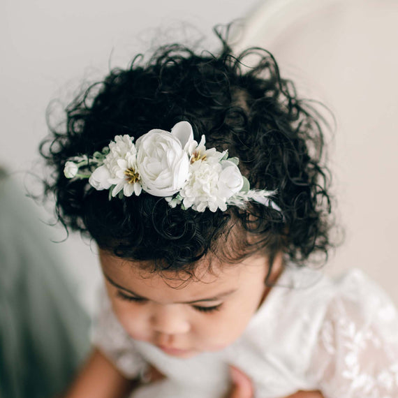 A baby wearing an Ella Flower Headband with delicate white flowers gazes downward. The baby has curly dark hair and is dressed in the Ella Romper Dress.
