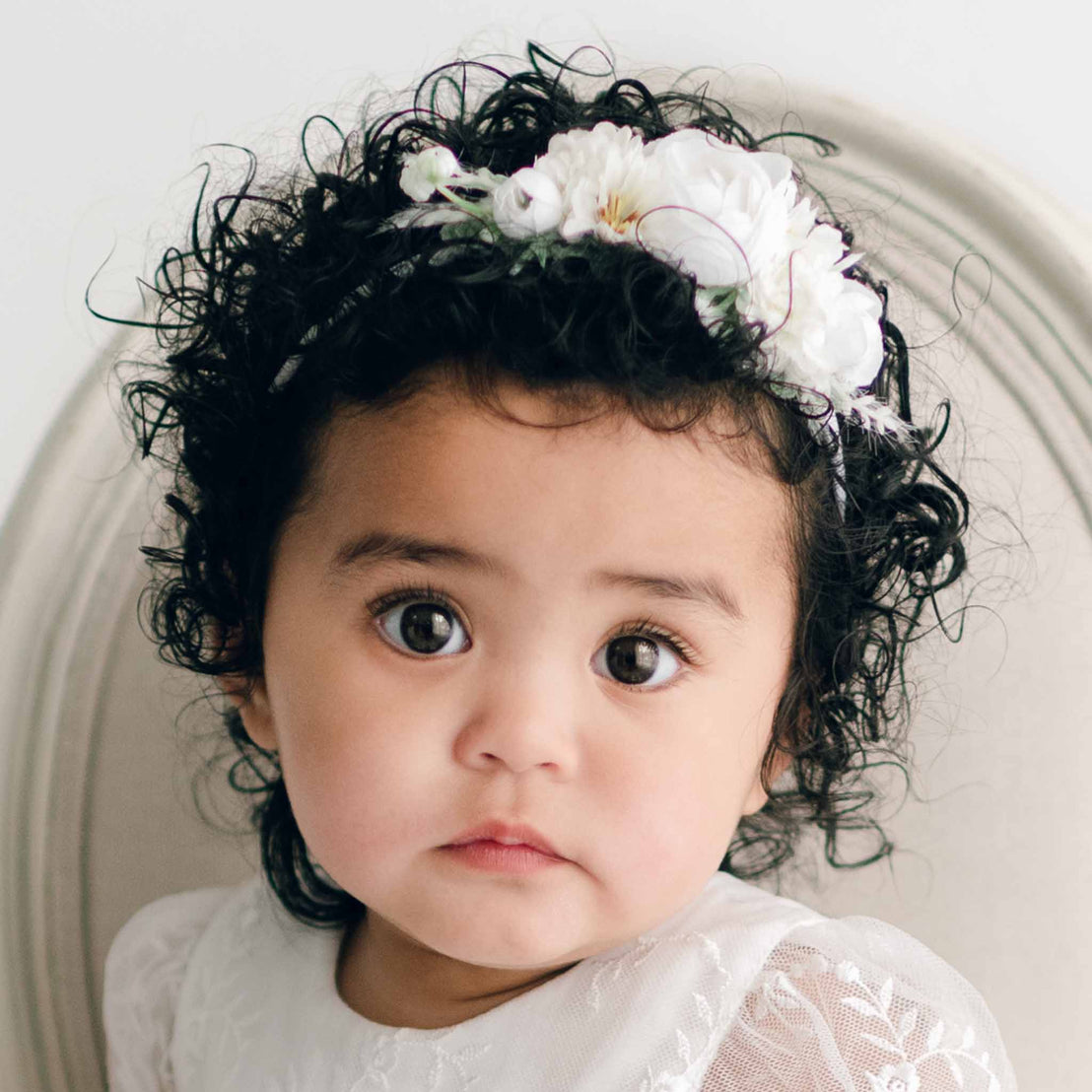 A baby with curly dark hair adorned with the Ella Flower Headband featuring white flowers sits against a neutral-colored background. Their expression is calm and curious.