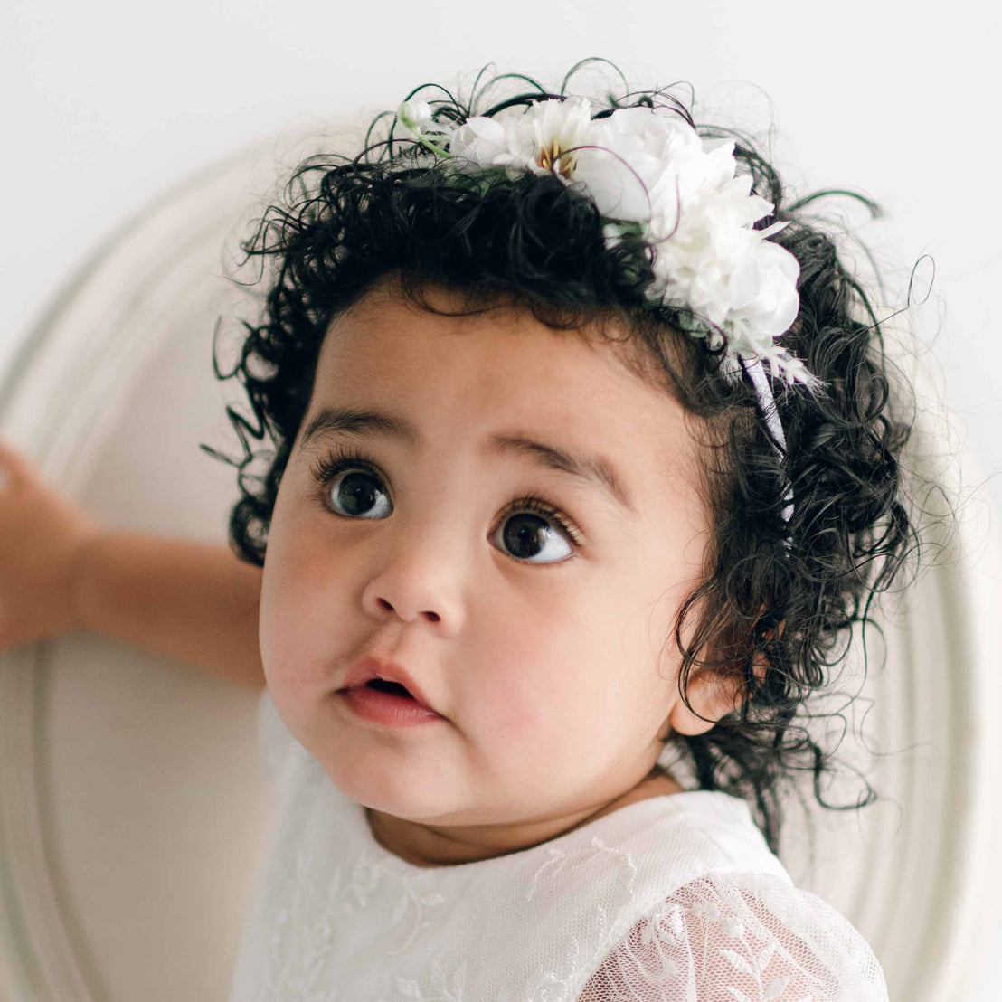 A baby girl with curly hair looks slightly upward with a curious expression. The child wears the Ella Romper Dress and an Ella Flower Headband made of white flowers. The girl rests one hand on a piece of furniture.
