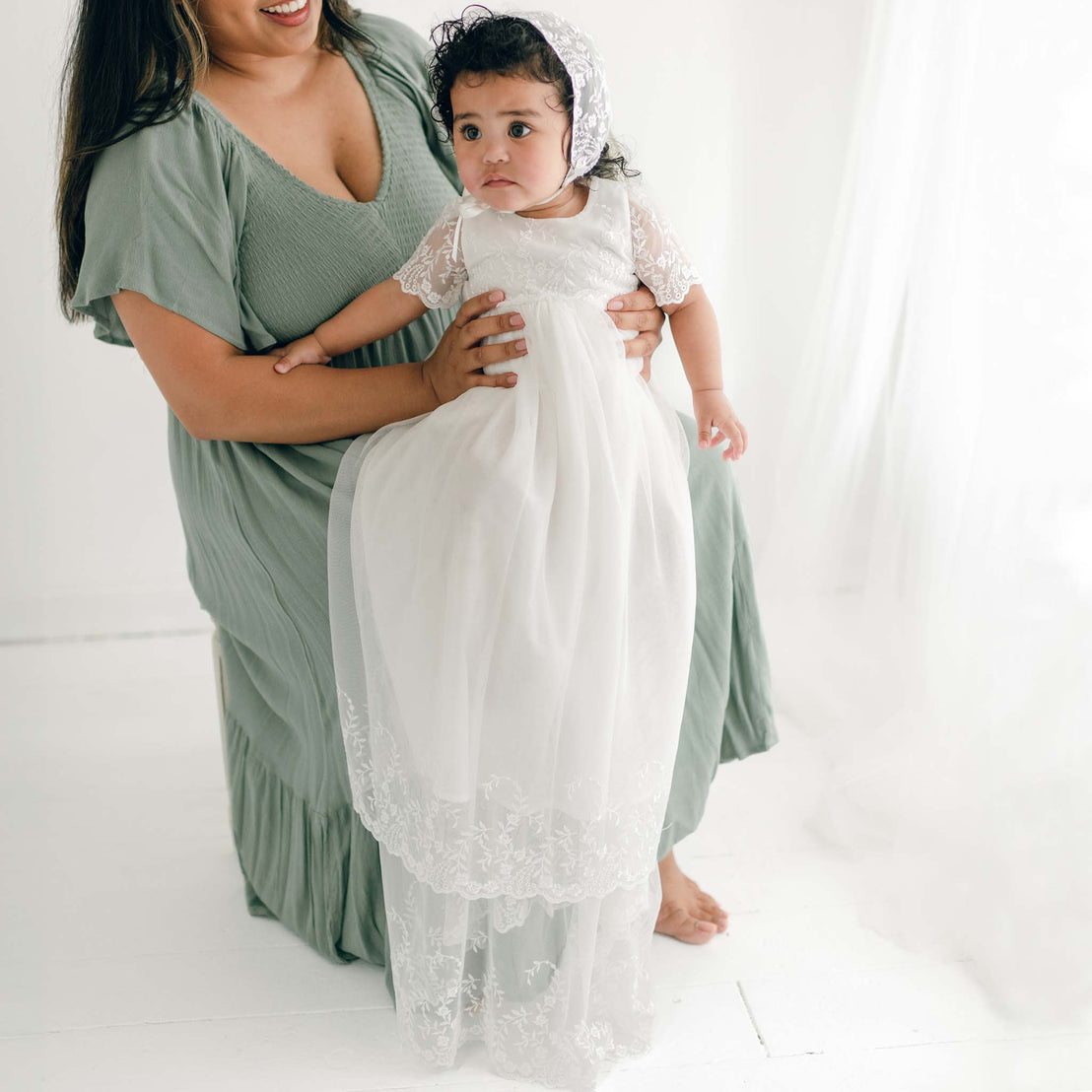 A woman in a green dress holds a baby wearing the Ella Christening Gown. The setting is a bright room with sheer white curtains. The woman is seated on a chair, smiling at the baby.