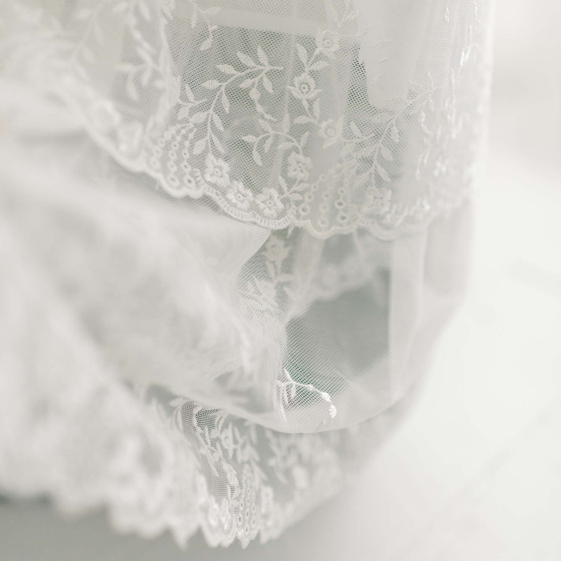 Close-up image of delicate, white lace fabric with intricate floral patterns and scalloped edges, featured on the Ella Christening Gown. The background is softly blurred.