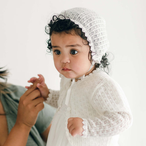 A young child with curly hair wears a white knitted bonnet and a matching Ella Knit Sweater, looking off to the side. An adult hand is gently holding the child's hand, and the background is plain white. The child has a thoughtful expression.