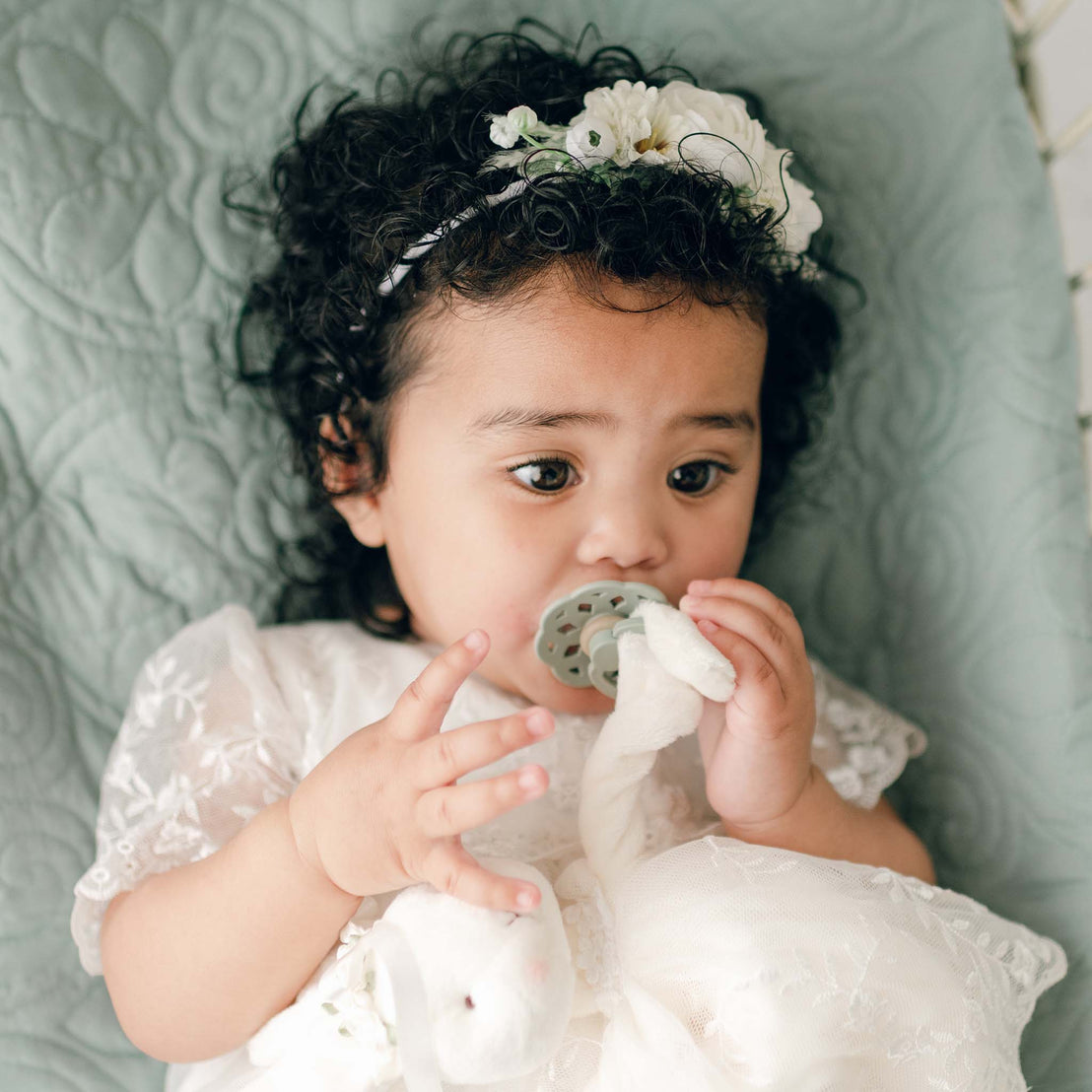 A baby with curly hair and wearing the Ella Flower Headband lies on a quilted surface. The baby holds an Ella Pacifier in sage and an Ella Silly Bunny Buddy, appearing engaged and curious.