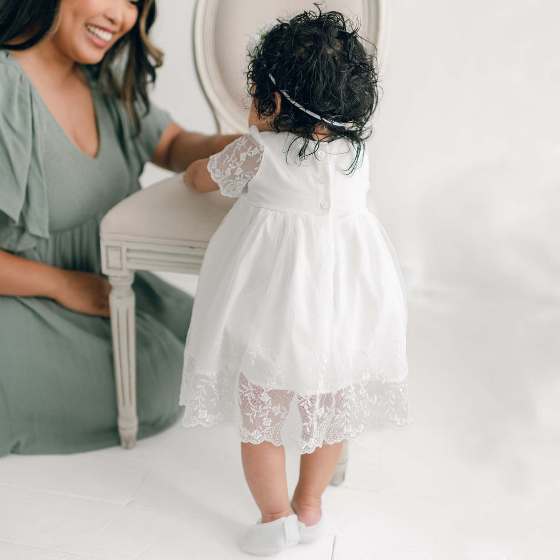 A smiling woman sits beside a toddler in a white Vivienne Romper Dress, who stands next to a white bench. The room has a bright, soft background.
