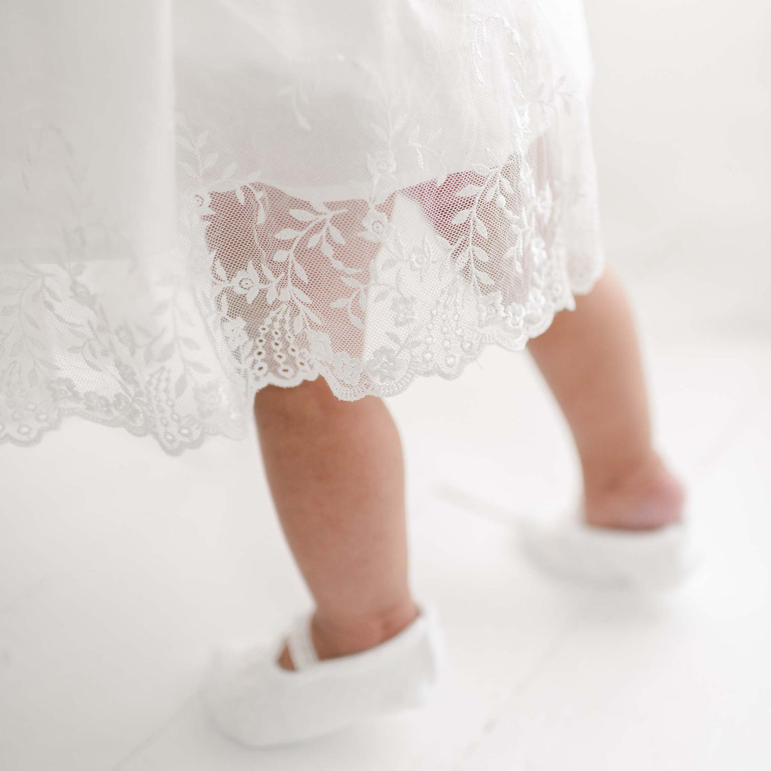 Close-up of a toddler's legs walking in white shoes and an Ella Romper Dress with floral embroidered netting lace on a white floor, capturing a moment of delicate movement.