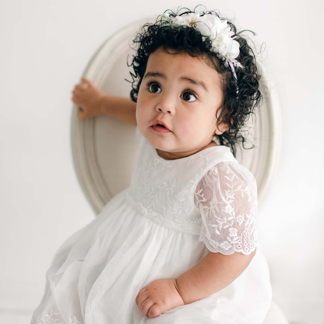 A toddler with curly hair and a floral headband wears a delicate Lilly romper dress made of floral embroidered netting lace, sitting in front of a round white backdrop, looking upwards with wide eyes.