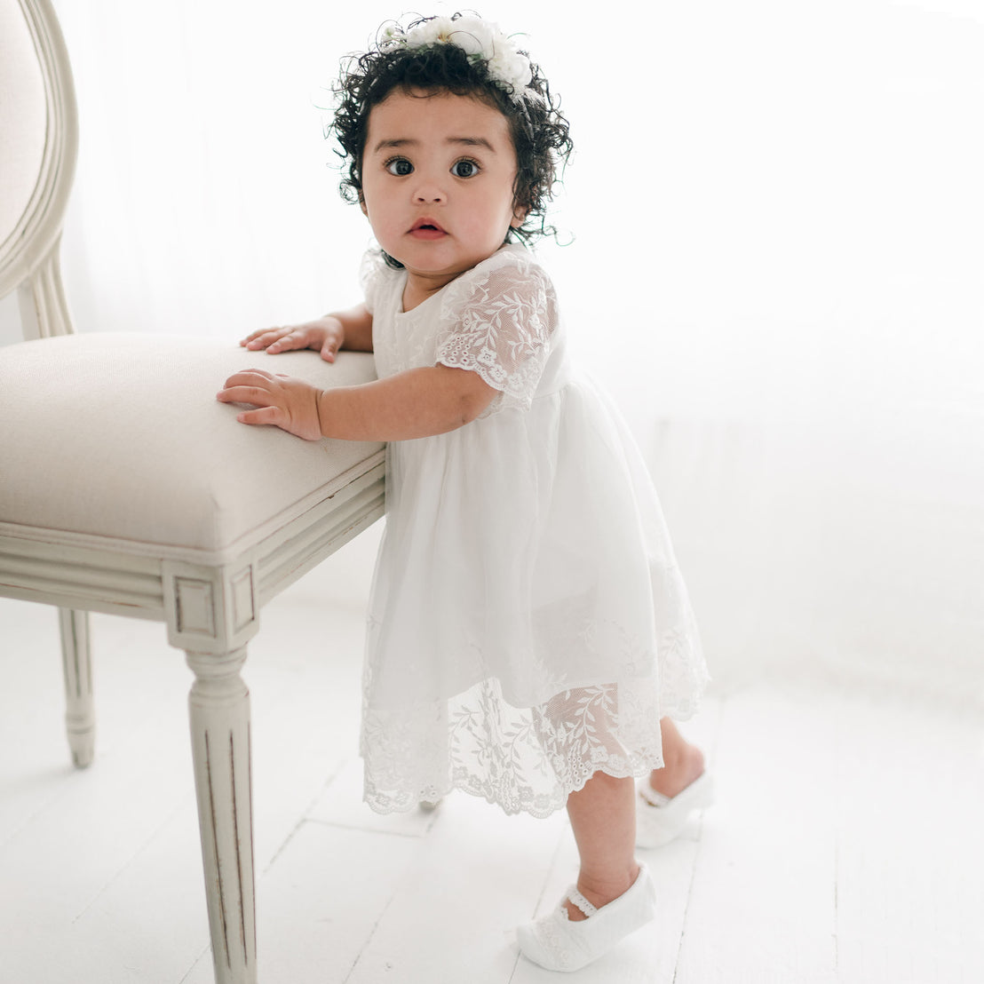 A toddler with curly hair, wearing an Ella Romper Dress and white shoes, stands and holds onto a cream-colored upholstered chair in a brightly lit room.