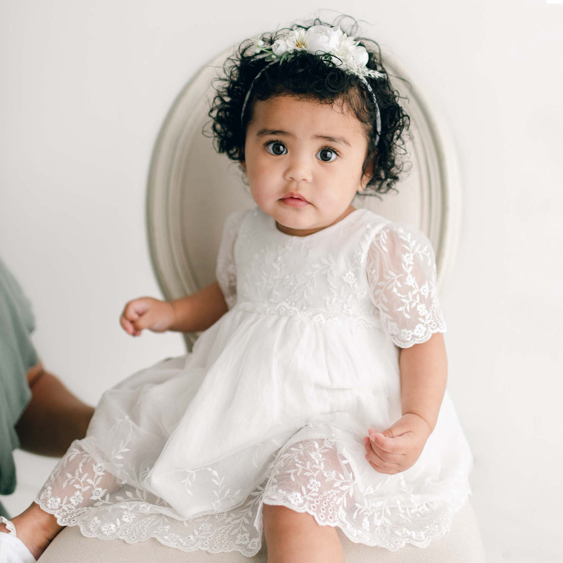 A baby girl with curly hair and big eyes, wearing an Ella Romper Dress with floral embroidered netting lace, sits on a vintage chair against a white backdrop.