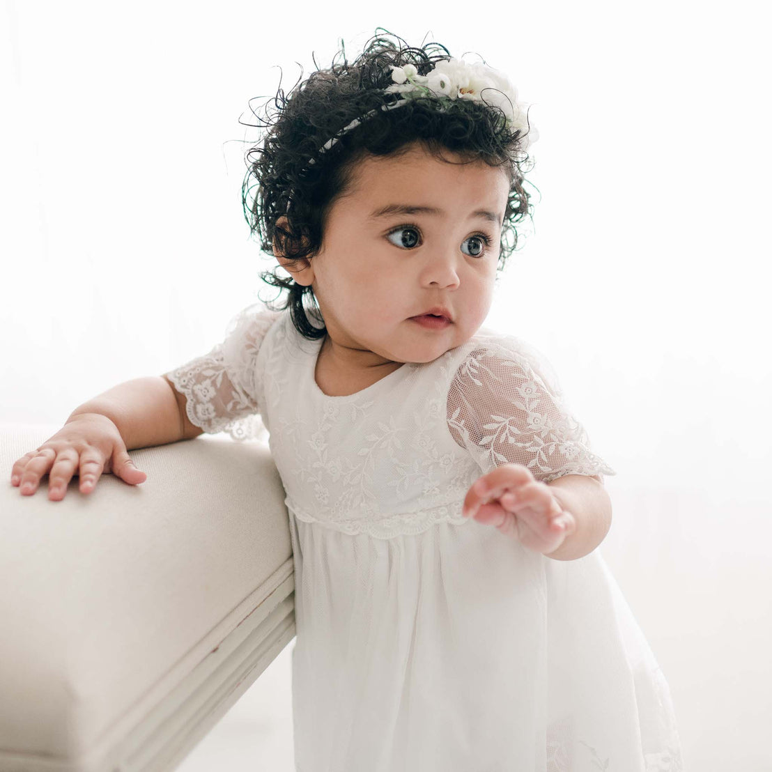 A toddler with curly hair wearing the Ella Romper Dress and a floral headband leans on a cream sofa, looking curiously to the side with wide eyes.