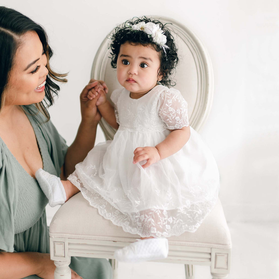 A woman smiles at a baby in an Ella Romper Dress sitting on an elegant chair. The baby wears a floral headband and looks curiously towards the camera. The background is bright and soft-focused.