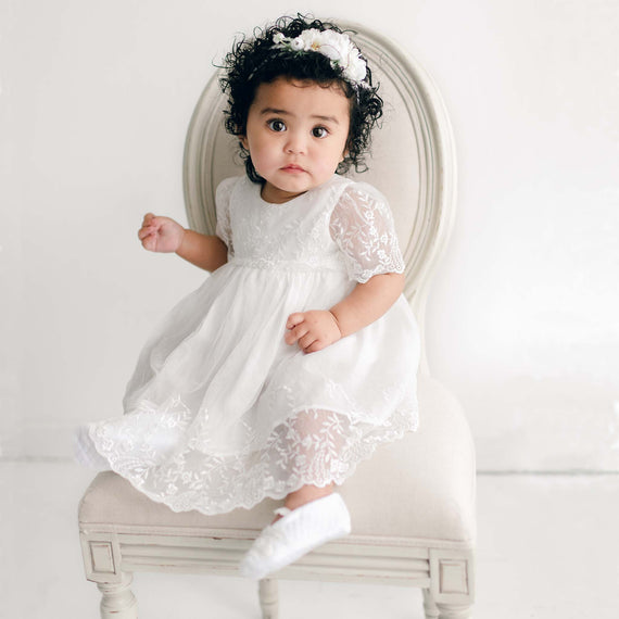 A toddler with curly hair wearing the Ella Romper Dress and a headband sits on an elegant chair, looking slightly to the side with a curious expression.