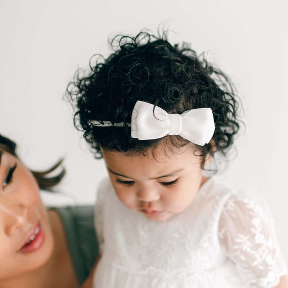 A mother softly looks at her young daughter, who is dressed in a delicate white lace dress with an Ella Velvet Bow Headband in her curly hair. They share a tender moment in a light, airy setting.