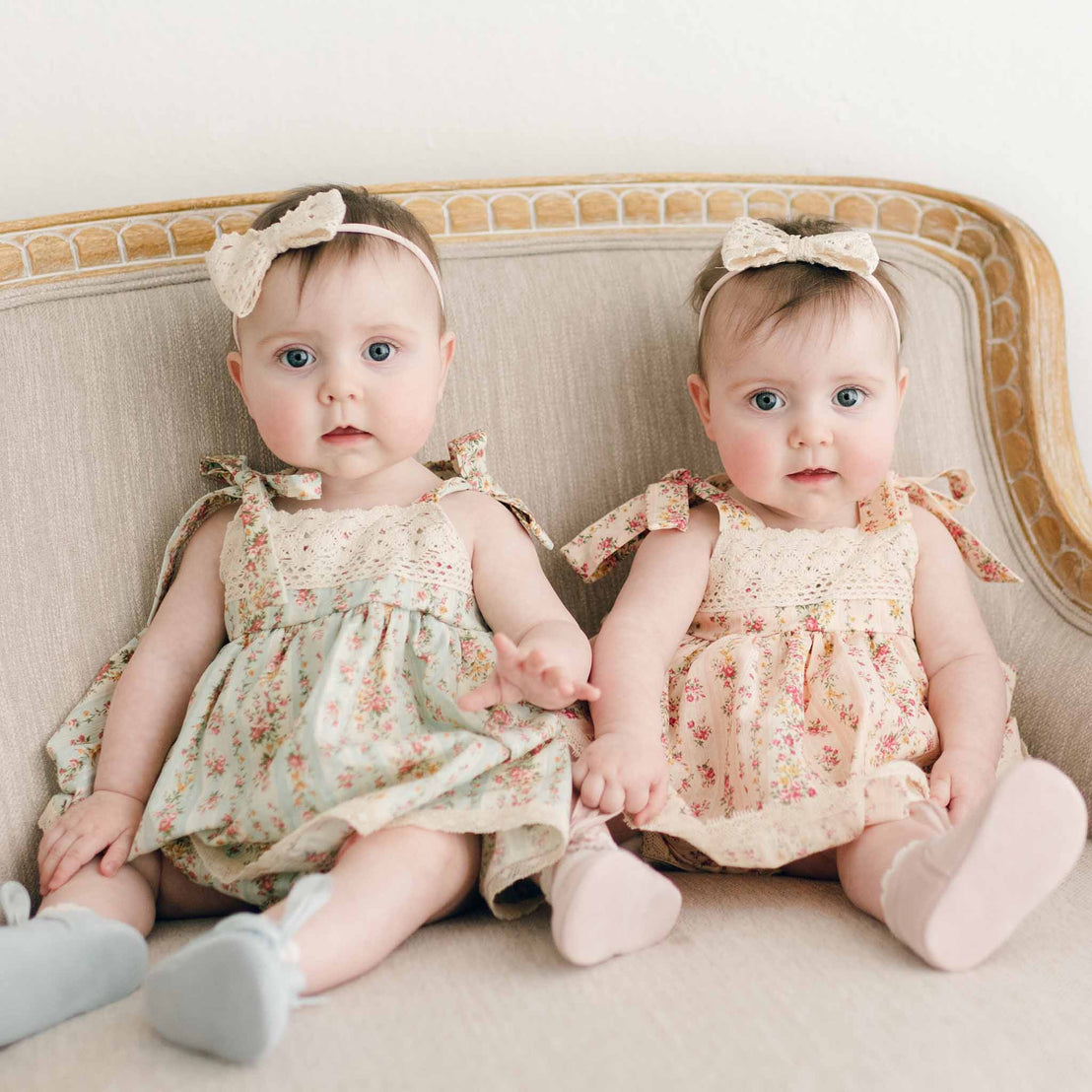 Two twin baby girls wearing the Eloise Romper Dress. One in Powder Blue and the other in Blush. They are also wearing the Eloise Bow Headband.