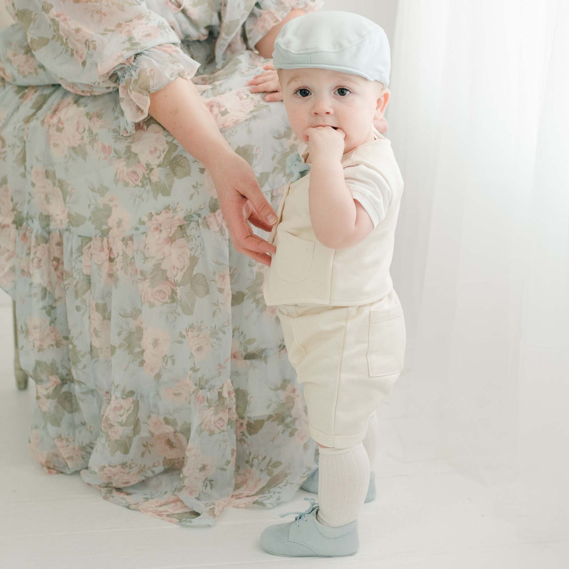 Baby boy standing near his mother. He is wearing a tan vest, tan pants and a powder blue Newsboy Cap, and blue velvet bow tie