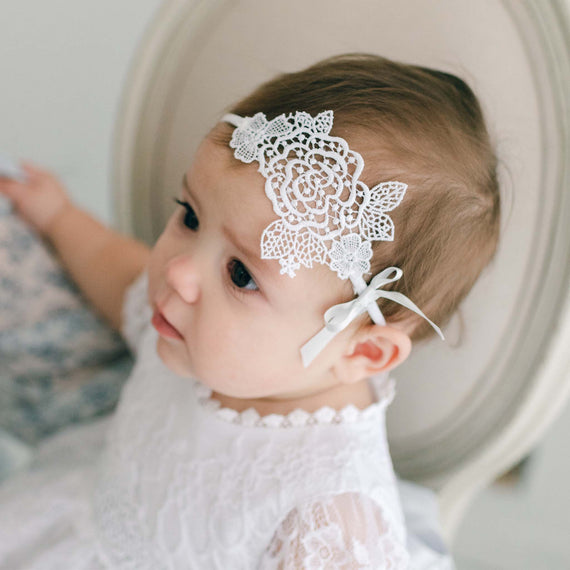 A baby wearing a white lace dress and the Olivia Lace Headband looks to the side, seated against a soft background, perfect as a boutique heirloom baby gift.