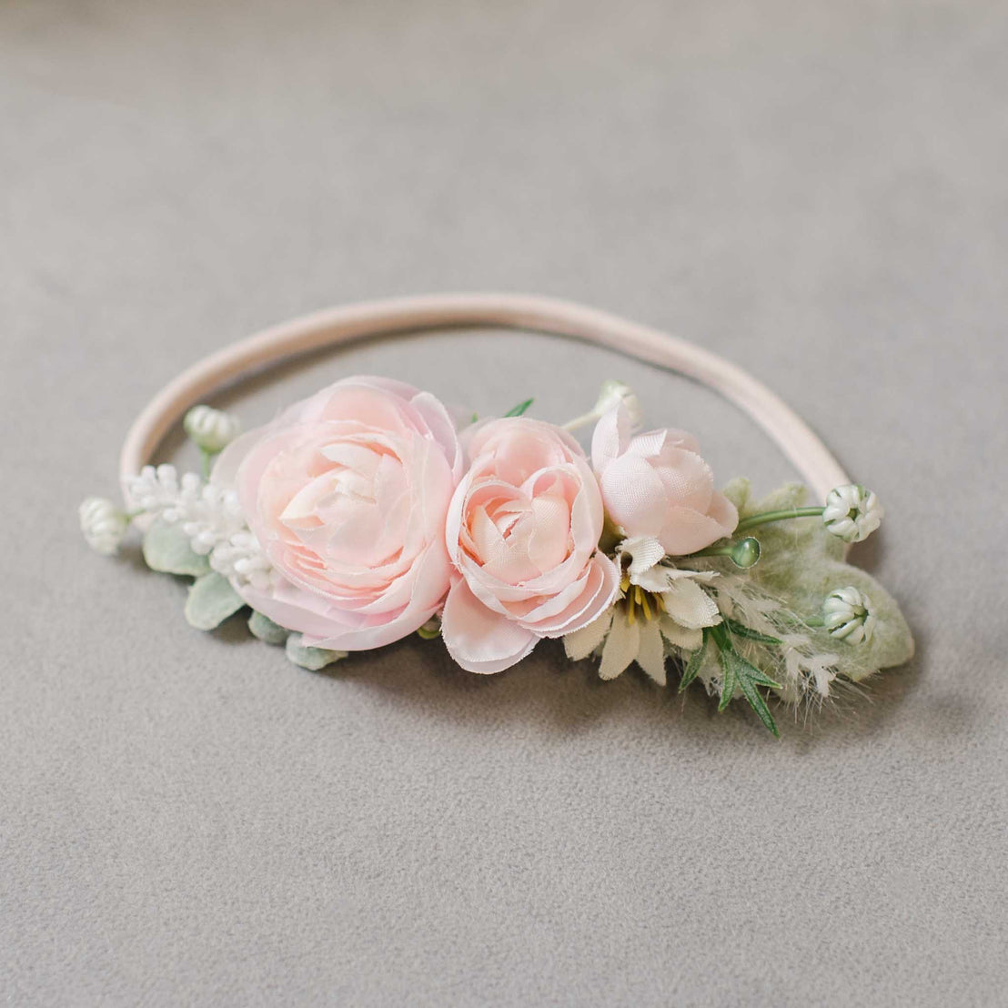 A delicate Joli Flower Headband featuring soft pink roses and white blossoms on a pale grey background, perfect for a boutique baptism.