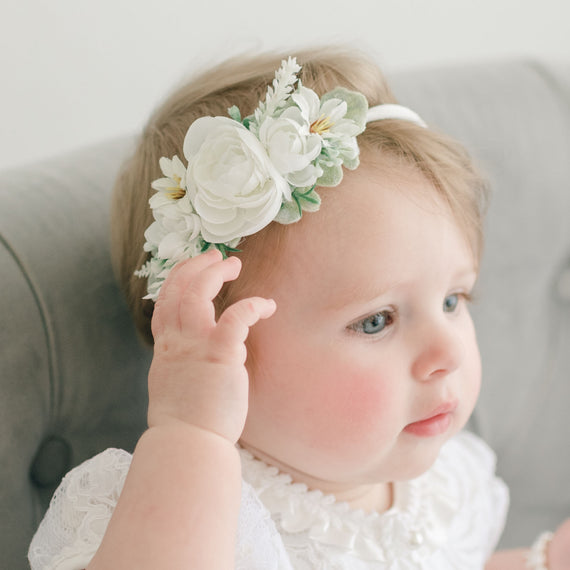 A baby girl with blue eyes and light hair wearing the Aria Christening Gown and the Aria Flower Headband, gently touching the headband while sitting on a grey chair.