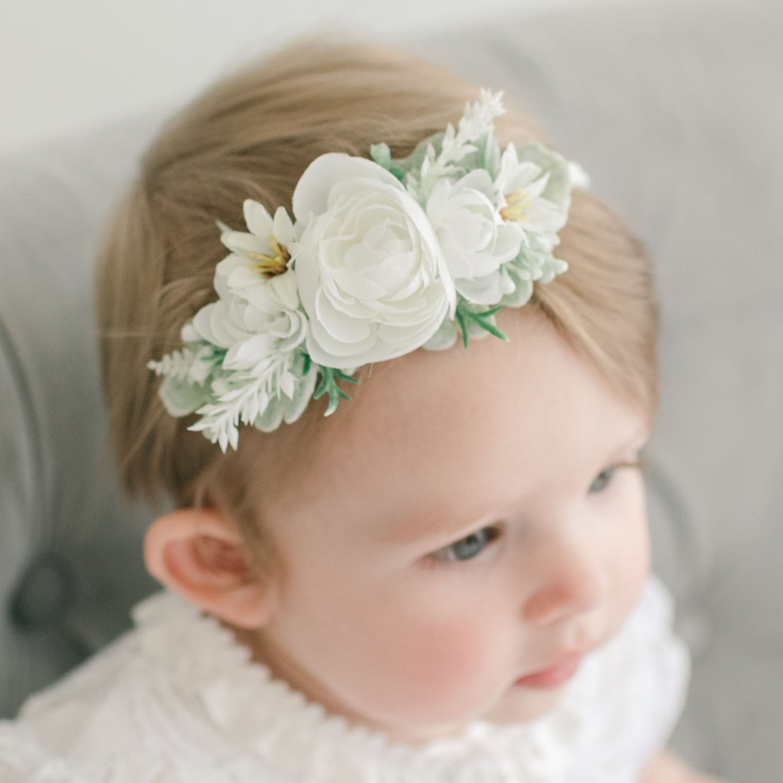 A close-up of a baby girl with light hair wearing the delicate handmade Aria Flower Headband, gazing off to the side. The child is dressed in the Aria Christening Gown.