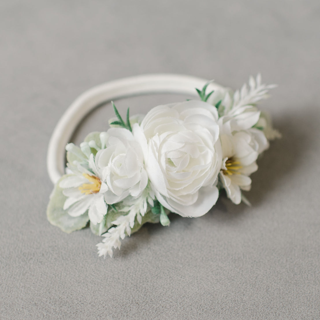The Aria Flower Headband that features a delicate floral arrangement of white flowers and green leaves on a super soft nylon band. The Aria Flower Headband lies on a textured grey background.