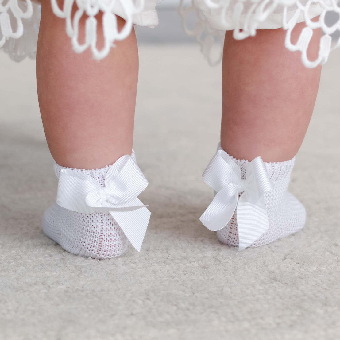 Close-up of a baby's feet in Garter Stitch Socks with Bow adorned with grosgrain ribbon bows, standing on a soft carpet.