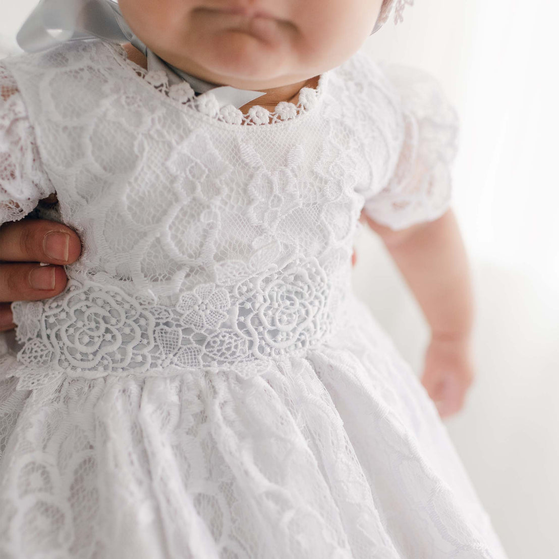 A close-up of an infant in an Olivia Christening Gown & Bonnet being held by an adult, with a focus on the detailed texture of the fabric and the baby's chubby arm.