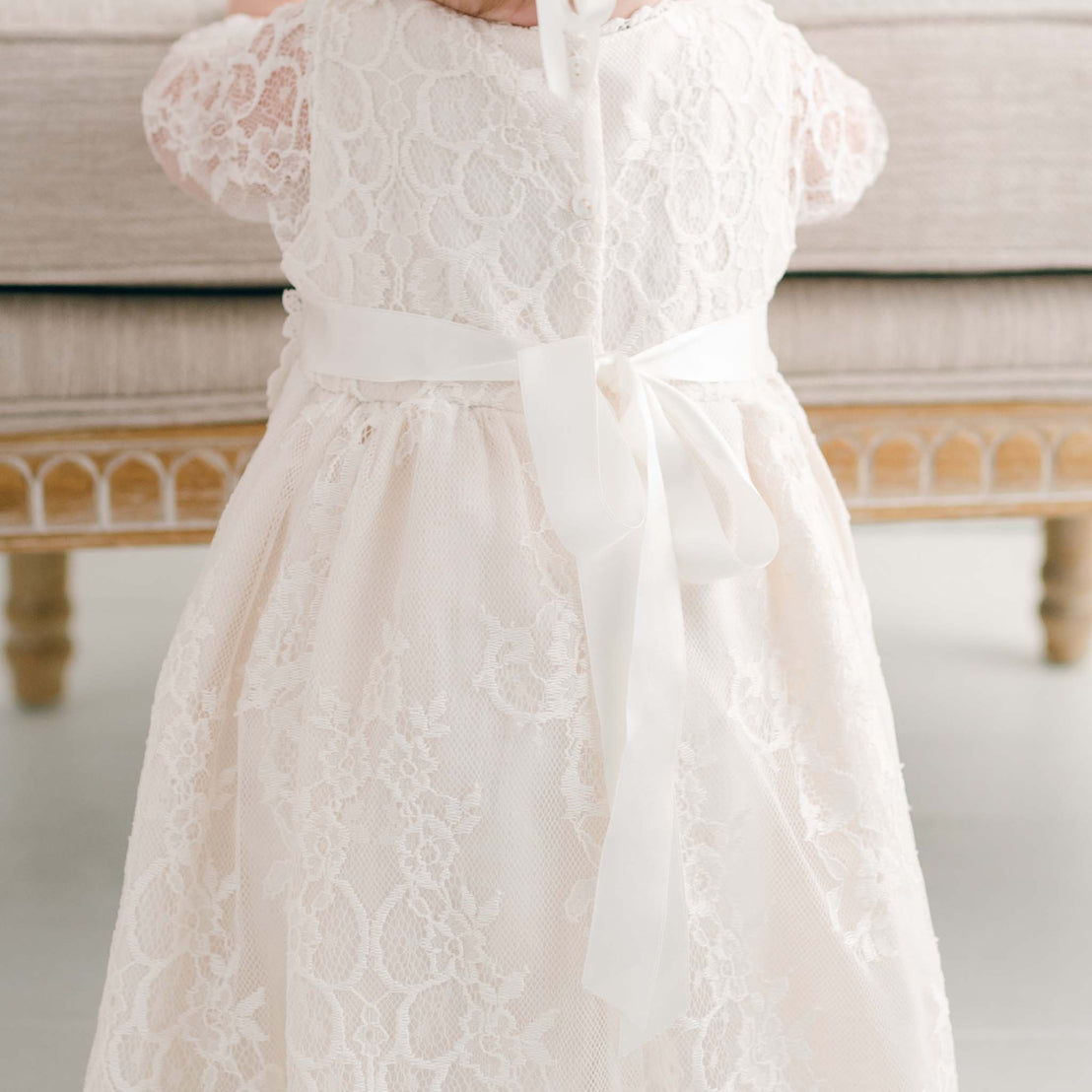 A close-up of a child wearing the Juliette Christening Gown & Bonnet, standing in front of a beige sofa. Only the torso and part of the arms are visible.