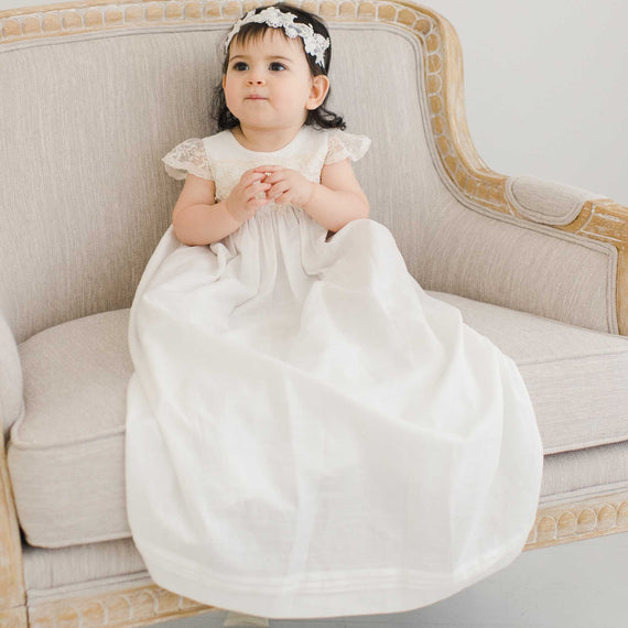 A toddler in a Jessica Linen Christening Gown & Beaded Flower Headband sits on a vintage sofa, her hands clasped together, looking thoughtful. The background is a soft, pale shade enhancing a serene atmosphere.
