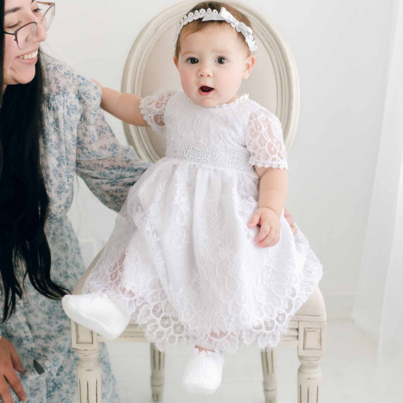A toddler in an Olivia Dress is held by her mother while sitting in an antique chair, both facing away from the camera in a softly lit boutique room.
