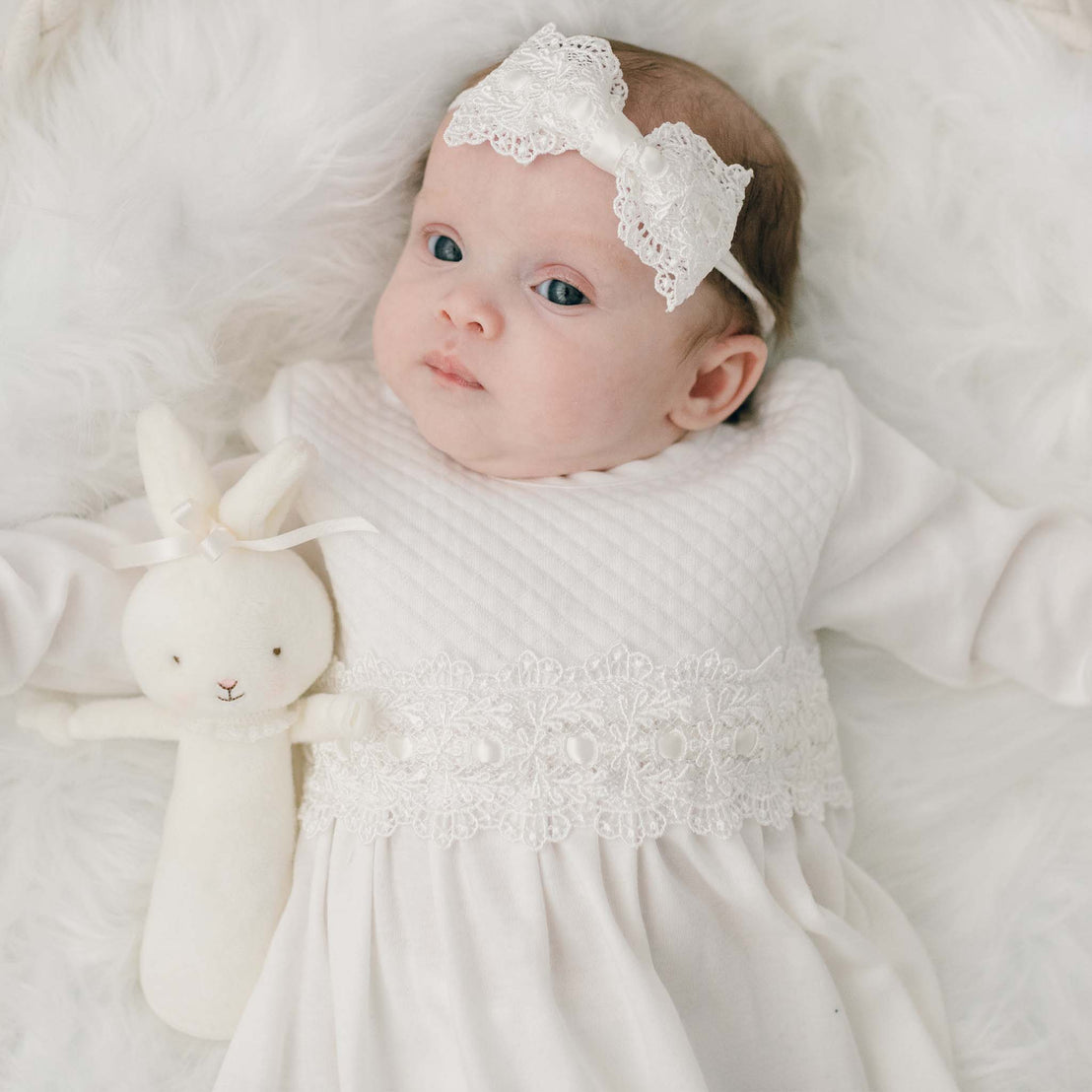 A newborn baby wearing a Madeline Newborn Gown lies on a furry blanket, gazing upward, holding a soft bunny toy, the perfect upscale baby gift from our boutique.