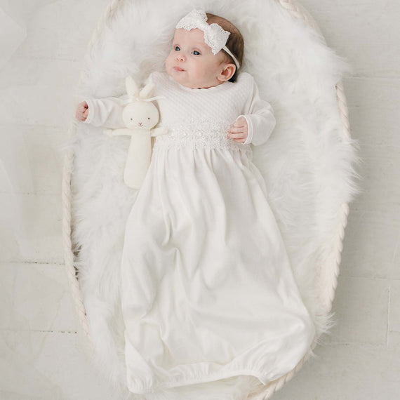 A baby with a headband lies in an upscale, cozy white basket, holding a stuffed rabbit, dressed in a Madeline Newborn Gown, on a fluffy rug.