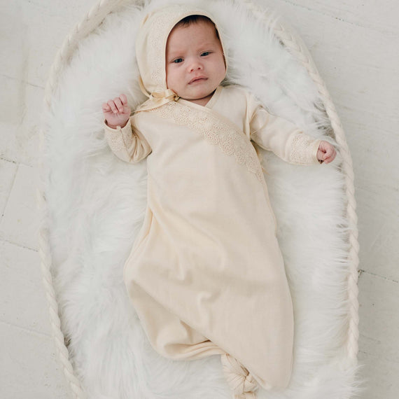 A newborn baby girl dressed in the Mia Knot Gown & Bonnet, a layette gown and matching bonnet made from a champagne pima cotton with embroidered lace and gold silk ribbon ties, lies on a white fluffy blanket.