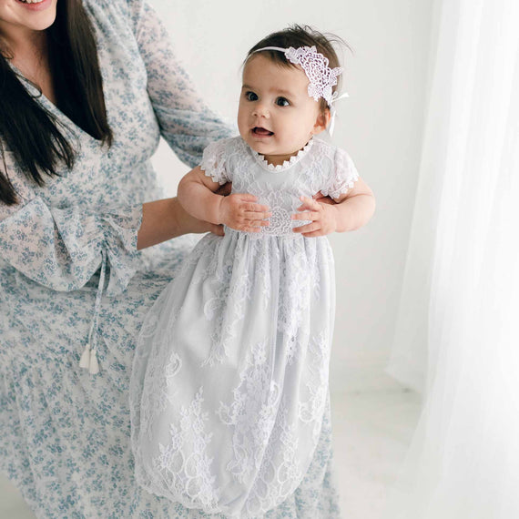 A baby in a delicate white lace Olivia Layette dress and headband, perfect for a baptism, is held by her mother wearing a floral dress. They are in a bright, softly-lit room.