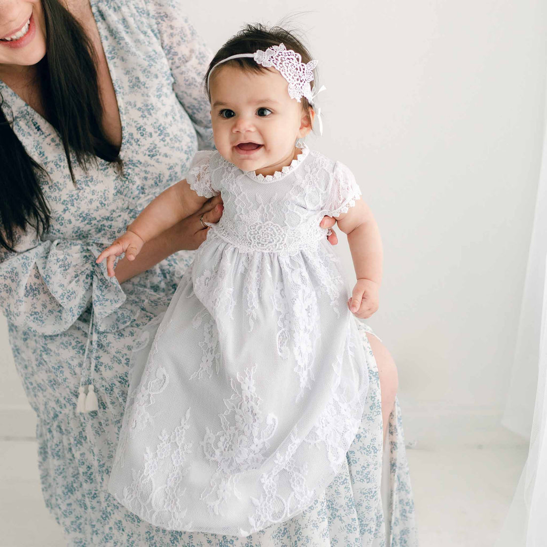 A smiling baby in an Olivia Layette and headband stands supported by a woman's hands, partially visible, in a floral dress against a white backdrop for a baptism.