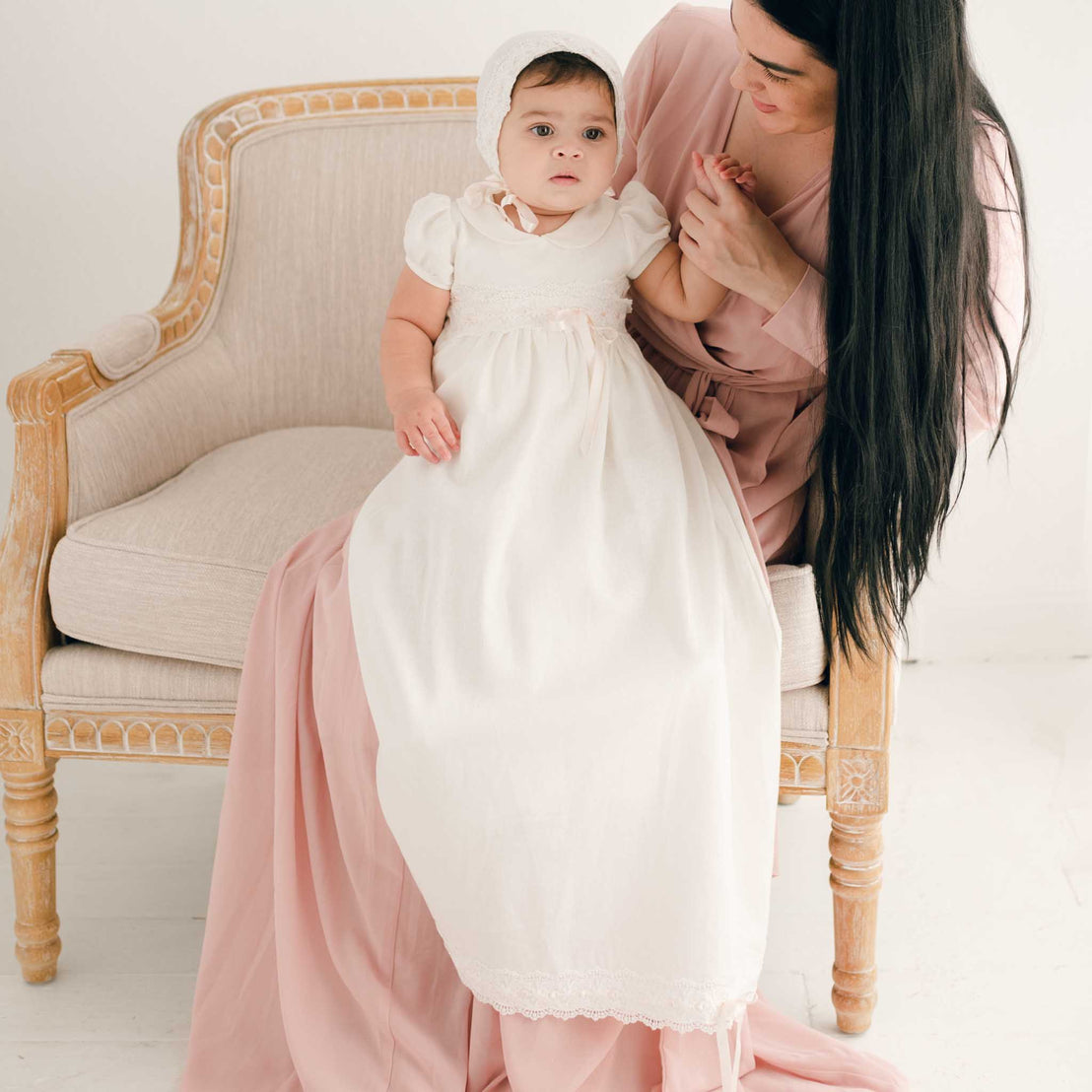 A woman comforts a baby in a long white gown sitting on an upscale beige armchair in a white room during a baptism.
