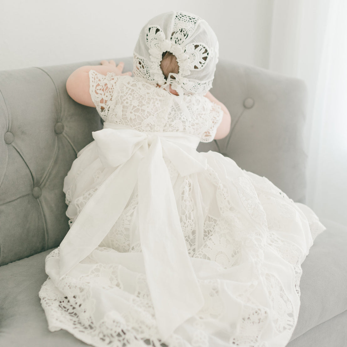 A baby in a Grace Christening Gown & Bonnet sits on a gray tufted couch, playful and content. The focus is on the intricate textures of the back of the dress.
