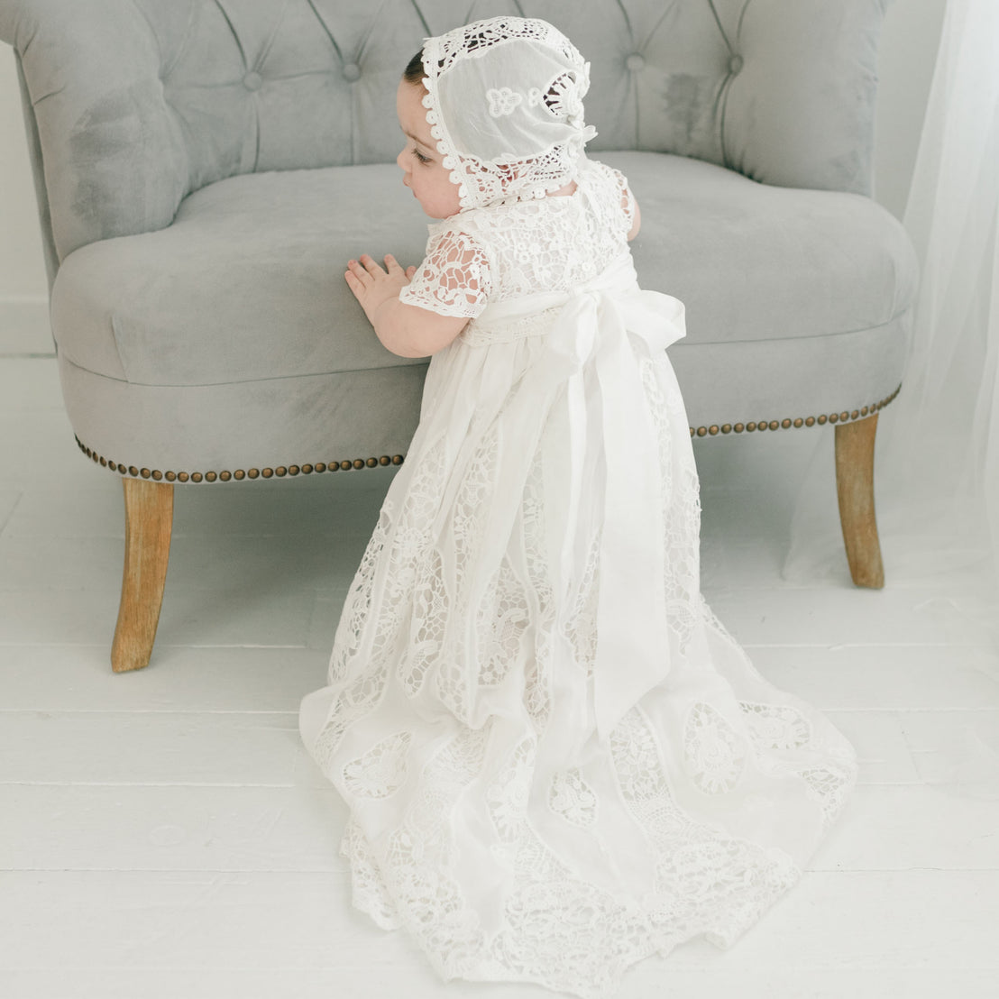 A baby wearing the Grace Christening Gown & Bonnet with a silk sash tie and Venice inset leans on a gray vintage sofa in a bright room.