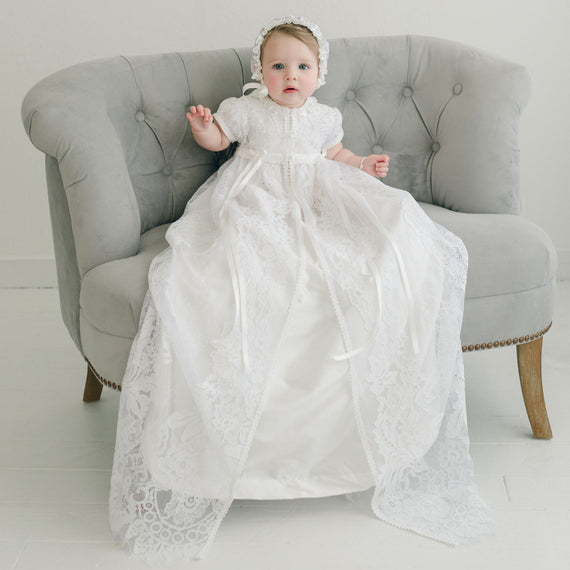 A baby wearing the Aria Christening Gown & Bonnet sitting on a gray armchair, looking directly at the camera with a neutral expression.