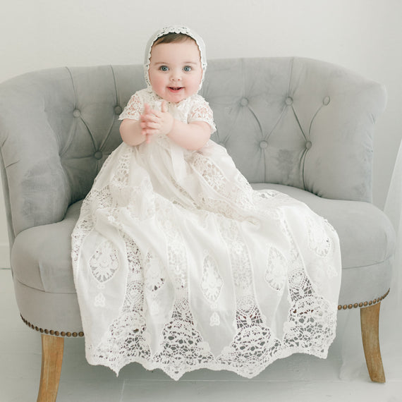 A joyful baby in a Grace Christening Gown & Bonnet with a Venice inset sits on a gray tufted sofa, smiling at the camera. The baby wears a matching lace cap.