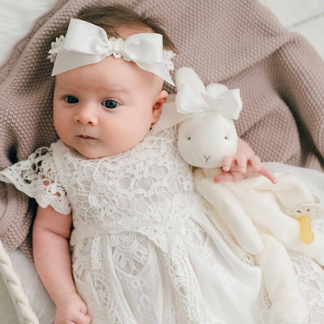A baby wearing a white Grace Dress and a white bow headband, lying on a soft blanket and holding a plush bunny toy.