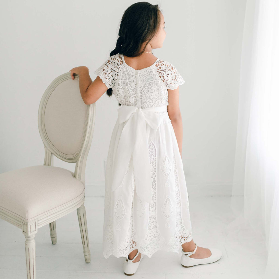 A young girl in a white Grace Girls Lace Dress stands next to a beige chair, looking away from the camera.