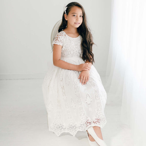 A young girl in a Grace Girls Lace Dress sits gracefully on a stool, her long hair flowing.