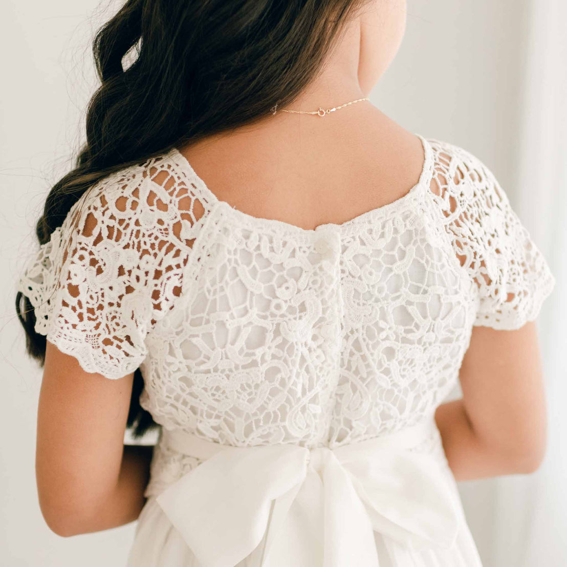 Girl photographed from behind, showcasing the detailed lace design of a white Grace Girls Lace First Communion Dress with short sleeves and a ribbon-tied waist.